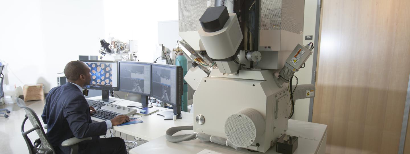 A person works at a computer with three screens displaying output from the FEI Helios 600 to the right of the workstation.