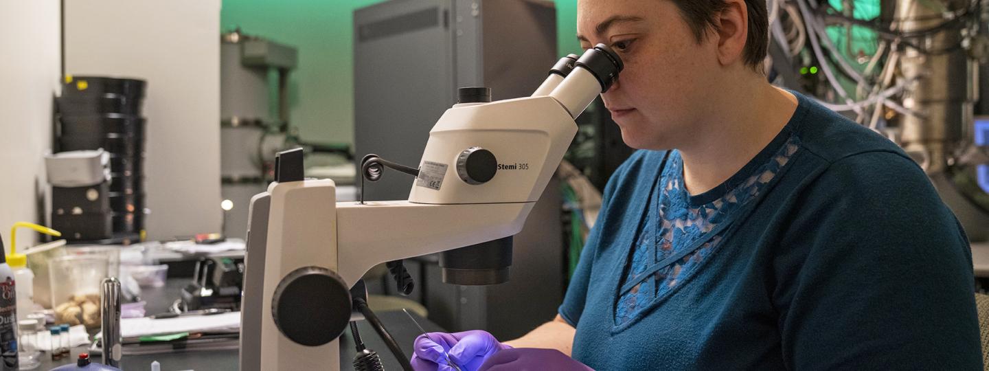 An individual looks into a microscope while preparing a sample.