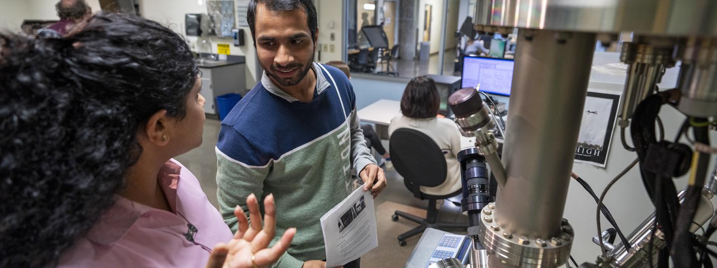 Two people talk in a lab in front of a large spherical machine while another researcher works at a monitor.