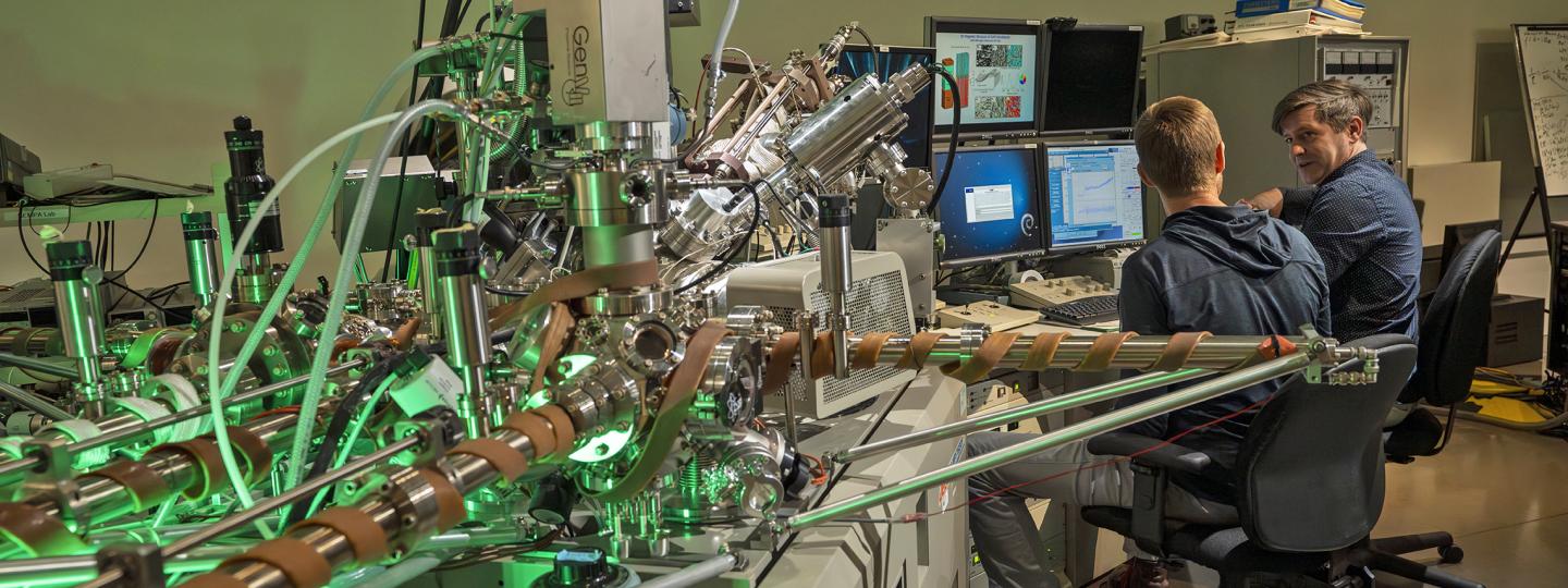 A very complicated large machine, made of shiny silver. Some of its parts are wrapped by wide brown tubing. Beside it, two people work a desk.