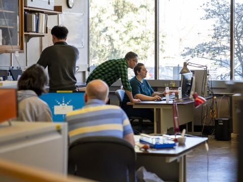 A busy lab interior. Four people work at two workstations with monitors, while one person works alone. Behind them are large windows with views of trees.