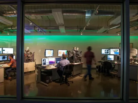 A person sits at a desk looking at one of multiple computer screens in front of them while another person walks behind them toward another workstation.