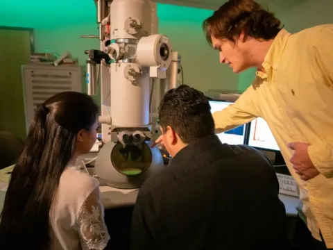 Thee people lean over an electron microscope.