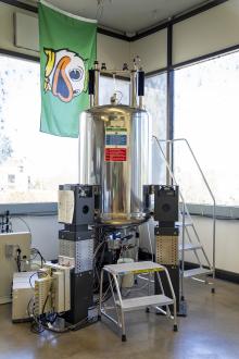 A large cylindrical machine stands next to two stepladders. The UO flag hangs in the background. 